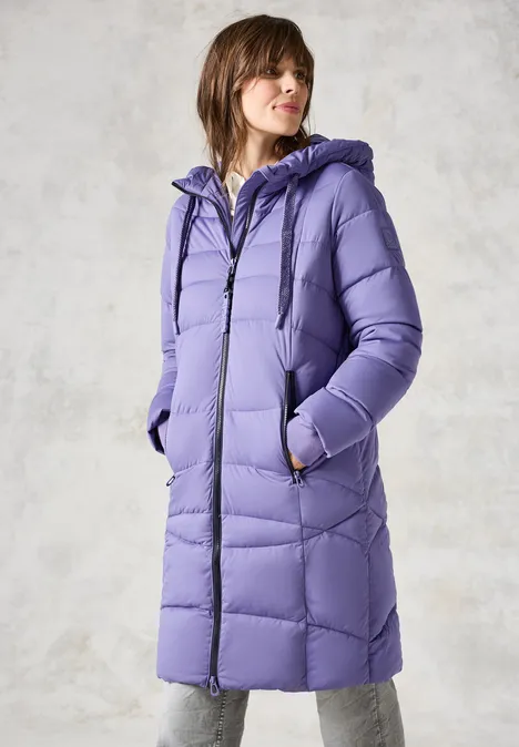 Frau in lilafarbener Steppjacke mit Kapuze vor grauem Hintergrund.